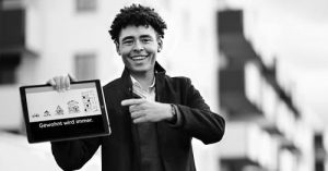 Junger Mann mit lockigem Haar hält ein Tablet mit der Aufschrift 'Gewohnt wird immer' in der Hand und lächelt. Hintergrund zeigt unscharfe Wohngebäude.